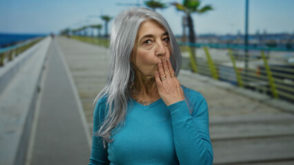 Senior woman with grey hair blowing a kiss on seaside promenade, capturing a serene outdoor beach...