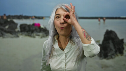 Senior woman making ok gesture in front of eye standing on a scenic seaside beach with grey hair...