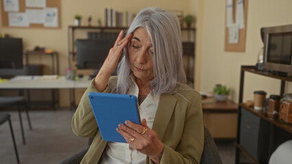 Senior woman with grey hair using a tablet in an office setting, exhibiting focus and concentration, seated amidst a cozy, well-organized workplace interior with decor and plants.