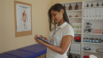 Young woman working with tablet in a medical clinic room showing medical chart and equipment in a...