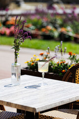 Summer cocktail on outdoor cafe table with flowers
Elegant cocktail in a crystal glass with herb garnish, placed on a sunlit cafe table surrounded by colorful blooming garden flowers.  
