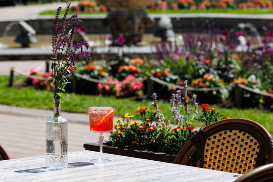 Refreshing cocktail on garden cafe table outdoors, A bright cocktail in a crystal glass placed on a cafe table with flowers and fountain in a colorful garden setting.
