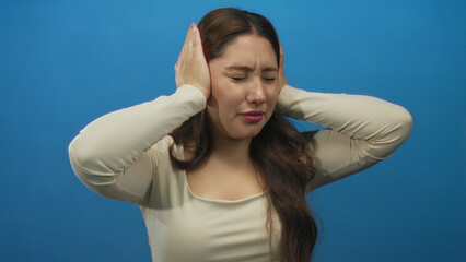 Young hispanic woman covering ears with hands and closing eyes against a bright blue studio...