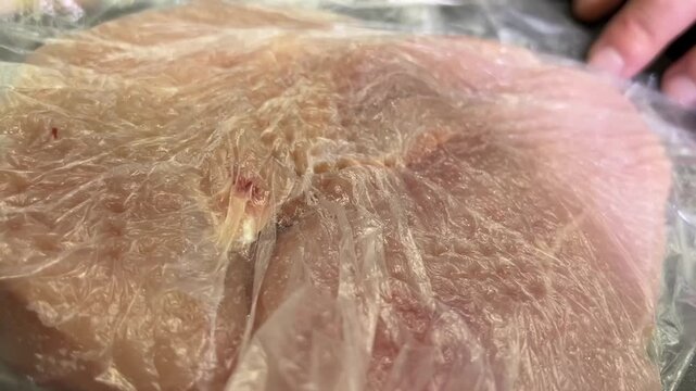 A hand pounds chicken breast under plastic wrap using a metal meat tenderizer on a kitchen counter