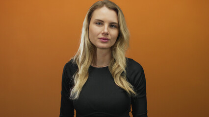 Woman with blonde hair in black shirt standing against a vibrant orange background, looking...