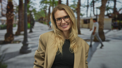 Woman smiling on a city street with palm trees wearing glasses and a beige jacket in an urban...