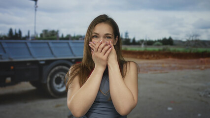 Young caucasian woman covers mouth with hands in front of truck on busy street under overcast sky;...