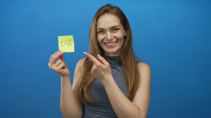 Smiling young caucasian woman holds yellow sticky note labeled tax with palm up against a blue...