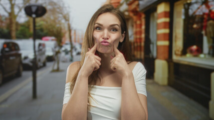 Young caucasian woman in white off shoulder top pinches cheeks and pouts on city street near brick...