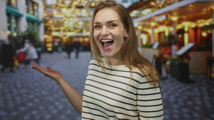 Young woman in striped top smiling and pointing finger to side on busy street under glowing cafe...