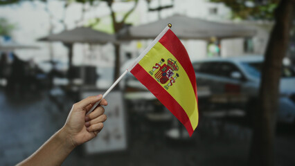 Hand holding spanish flag on outdoor cafe terrace in city with caucasian man in background creating...
