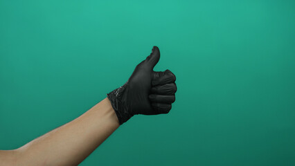 Caucasian male hand gives a thumbs up wearing a black glove against a solid green background...