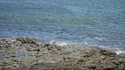Mediterranean seaside scene showcasing rocky shore with clear blue waves under a bright sunny sky, capturing the essence of nature's tranquility and coastal beauty.