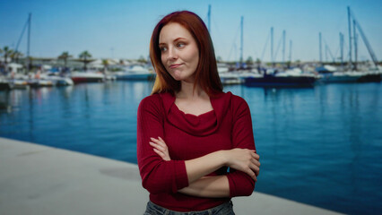 Woman with red hair stands confidently by the seaside with moored boats, showcasing beauty and...