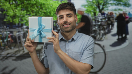Young hispanic man smiling while holding white gift box with blue ribbon on city street;...