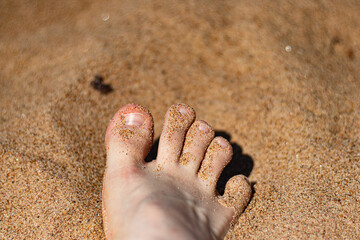 A close-up image of a bare foot partially buried in warm, golden beach sand. The foot is covered in fine grains, and the bright sunlight highlights the natural textures of both the skin and the sand. 