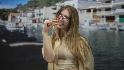 Fototapeta premium Woman with blonde hair pinches fingers near face at sunny seaside port with boats; tiny measurement.