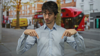 Fototapeta premium Man points both index fingers to light blue shirt on busy city street amid blurred red bus; confidence.