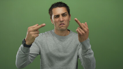 Young hispanic man shows double middle finger gesture in studio before green wall with...