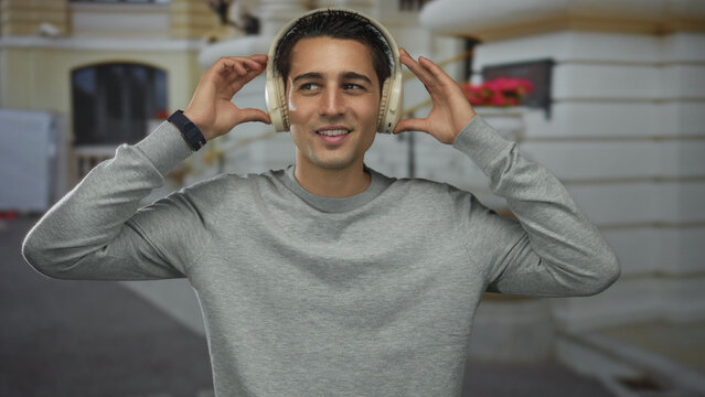 Young hispanic man enjoying music with headphones outdoors, smiling while wearing a casual sweater in front of a blurred urban backdrop, exuding a joyful and relaxed vibe. - Powered by Adobe