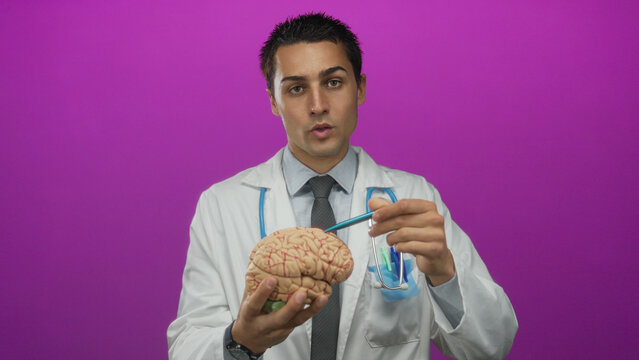 Young hispanic male doctor in uniform holding brain model with stethoscope around neck against isolated pink background