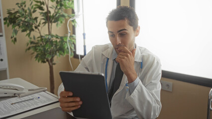 Young hispanic man doctor using tablet indoors hospital clinic office wearing uniform with stethoscope suggesting focused workplace environment.