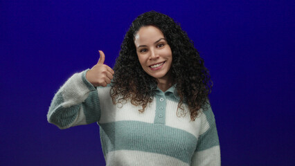 Smiling woman giving thumbs up over isolated blue background with curly hair wearing striped...