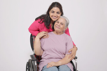 Indian Young woman helps and supporting to her senior mother suffering from knee pain. disabled senior mother and daughter embracing against grey studio background 