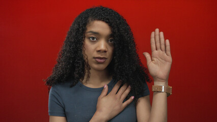 Woman with serious expression holding hand on chest and the other hand raised against red studio...