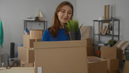 Woman in blue moving boxes in new apartment surrounded by packed items and smiling while holding...
