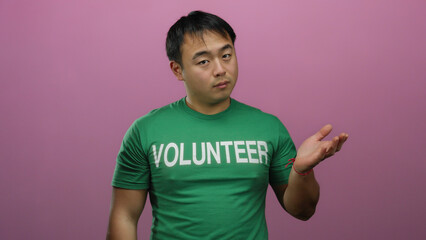 Young man wearing green volunteer shirt stands against isolated pink background in an expressive...