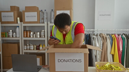 Asian man in a donation center checking items in a box labeled donations with a smartphone,...