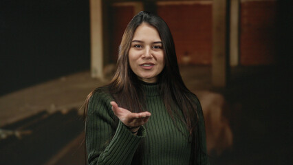 Woman sending kiss to camera stands in unfinished indoor construction site wearing green sweater,...