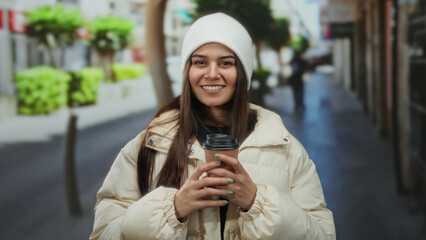 Obraz premium Woman enjoying coffee outdoors on a lively street, wearing a white coat and beanie, depicting a casual urban scene with a hispanic flair.