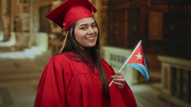 Woman celebrating graduation holds cuban flag in a church setting, wearing a red cap and gown indoor, symbolizing pride and achievement.