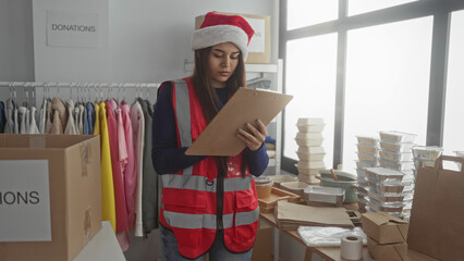 Woman wearing santa hat in charity center organizes donations on clipboard in indoor room...