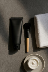Naklejka premium Minimal spa scene: black cosmetic tube, facial brush with wood handle, folded towel and swirl of cream on gray stone. Warm hard light, crisp shadows, clean unbranded setup.