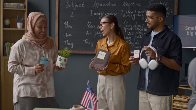 Medium shot of Caucasian female teacher and diverse group of students using sticky notes to label and describe plant, book, headphones, and other items during engaging English class