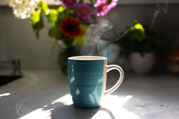 A steaming cup of coffee sits on a kitchen counter, illuminated by sunlight. Colorful flowers decorate the background, creating a warm and inviting atmosphere filled with morning energy.