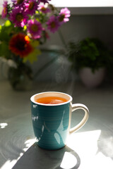 A turquoise ceramic mug filled with steaming tea sits on a light-colored table. Colorful flowers are arranged in the background, illuminated by soft morning light, adding warmth to the setting.