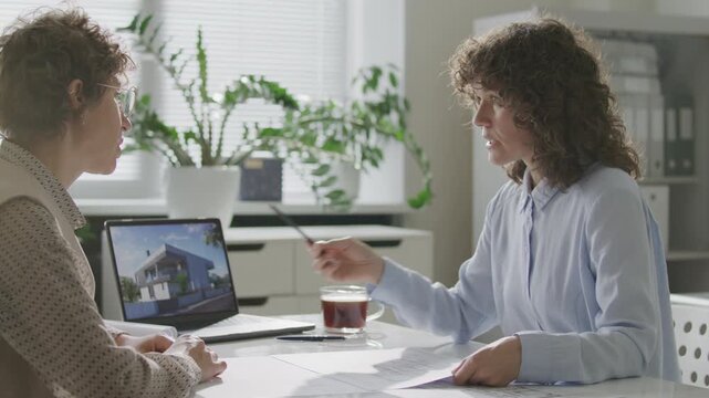 Female architect presenting modern building visualization on laptop and explaining floor plans to client during meeting in office