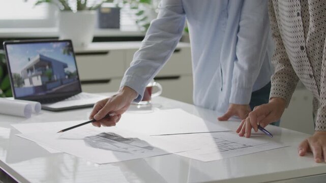 Two female colleagues examining printed architectural plans on office table during meeting, with laptop displaying house render nearby. Close-up view