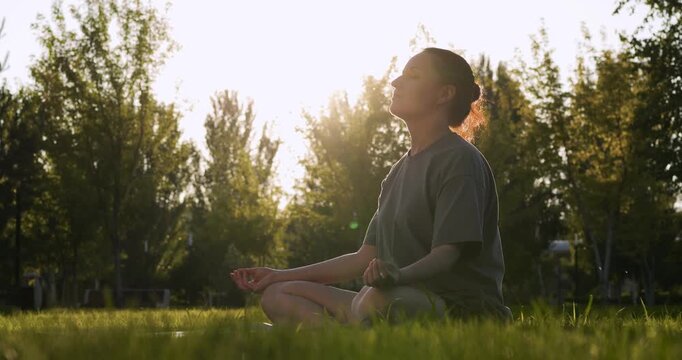 Woman meditates in lotus pose on grass in park at sunset during golden hour. Meditation in nature helps girl to find harmony balance. Meditation brings woman peace tranquility. Woman relaxes sunlight.