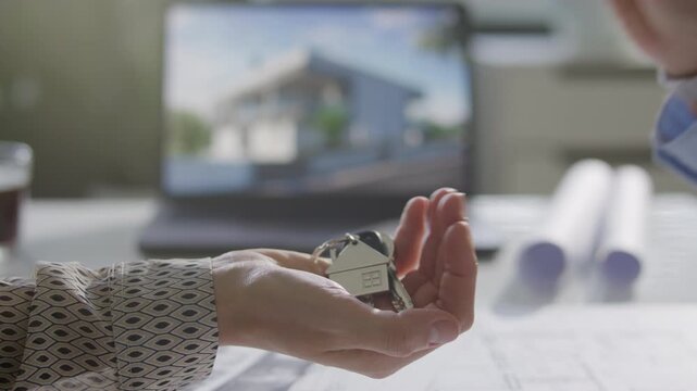 Close up view of hand of female property buyer receiving house shaped keychain with keys from seller over office desk during real estate handover