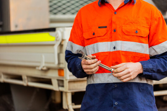 Young man in workwear standing behind vehicle holding a spanner in mechanics workshop