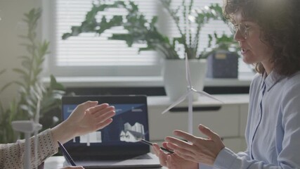 Two female colleagues reviewing presentation on laptop screen and discussing renewable energy project with wind turbine model on desk in office - Powered by Adobe