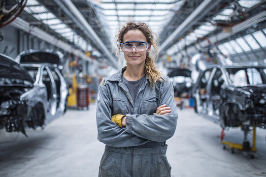 Proud female automotive worker in gray jumpsuit stands with arms crossed by a partially assembled car, wearing safety gear on the factory floor under daylight panels.