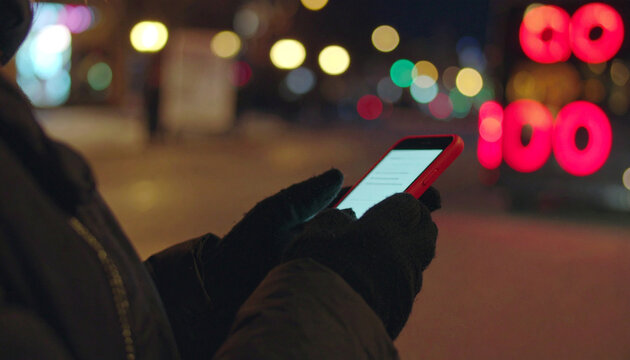 Digital Glow in the Night: A person's gloved hands holding a smartphone, illuminating the dimly lit street with its screen, blurring the vibrant city lights in the distance. - Powered by Adobe