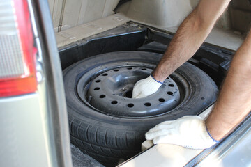 A brunette man in white work gloves takes a spare tire out of the trunk of a silver car.