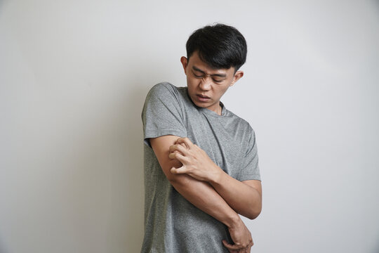 A young Asian man scratches his arm with a grimacing expression, indicating irritation or a skin problem.